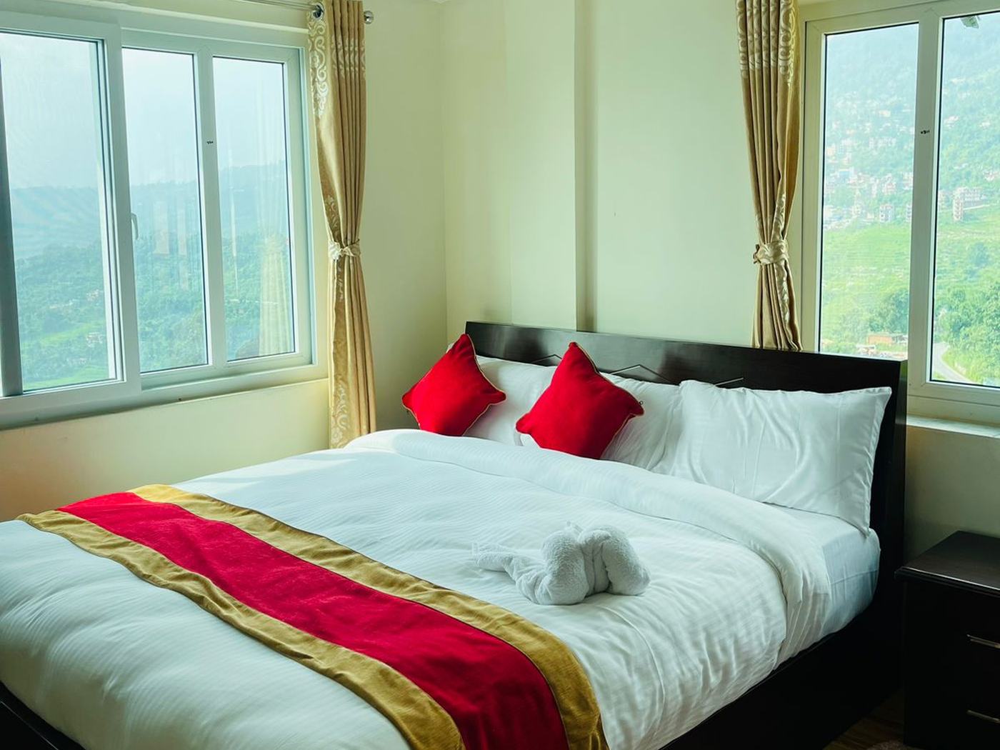 A double bed with white linen and a red and gold runner, between two windows looking out over the Gorkha hills
